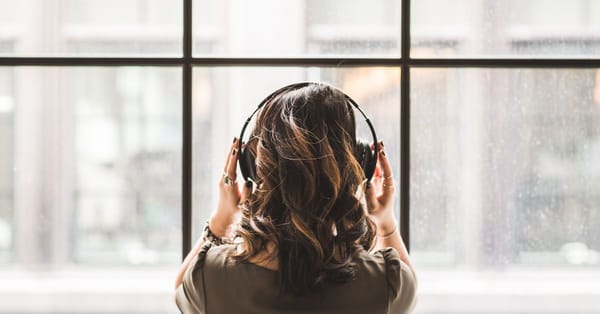 A woman looks out the window while putting over the ear headphones on.